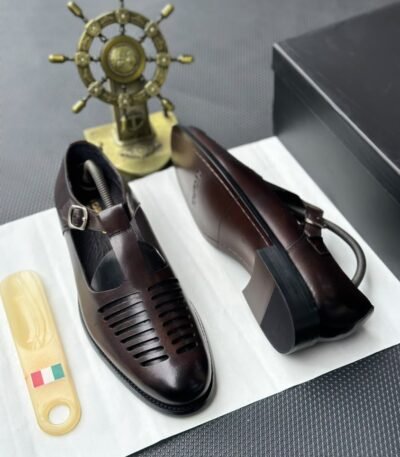 A pair of luxurious black leather shoes with a silver buckle and ventilation details, presented on a white cloth with a shoe tree and a decorative gold ship's wheel in the background.
