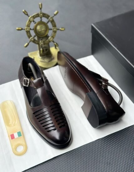 A pair of luxurious black leather shoes with a silver buckle and ventilation details, presented on a white cloth with a shoe tree and a decorative gold ship's wheel in the background.