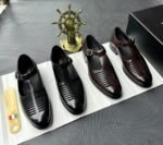 A pair of luxurious black leather shoes with a silver buckle and ventilation details, presented on a white cloth with a shoe tree and a decorative gold ship's wheel in the background.