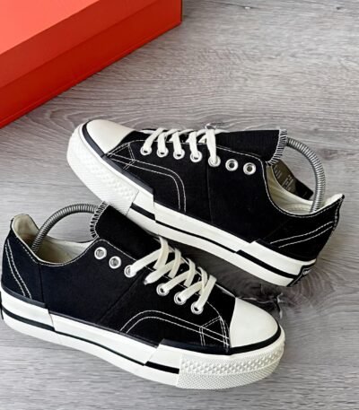 A pair of black sneakers with white laces and soles, placed on a wooden floor next to a red shoe box.