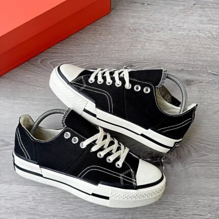 A pair of black sneakers with white laces and soles, placed on a wooden floor next to a red shoe box.
