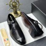 A pair of luxurious black leather shoes with a silver buckle and ventilation details, presented on a white cloth with a shoe tree and a decorative gold ship's wheel in the background.