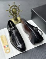 A pair of luxurious black leather shoes with a silver buckle and ventilation details, presented on a white cloth with a shoe tree and a decorative gold ship's wheel in the background.