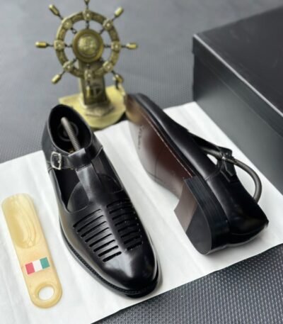 A pair of luxurious black leather shoes with a silver buckle and ventilation details, presented on a white cloth with a shoe tree and a decorative gold ship's wheel in the background.
