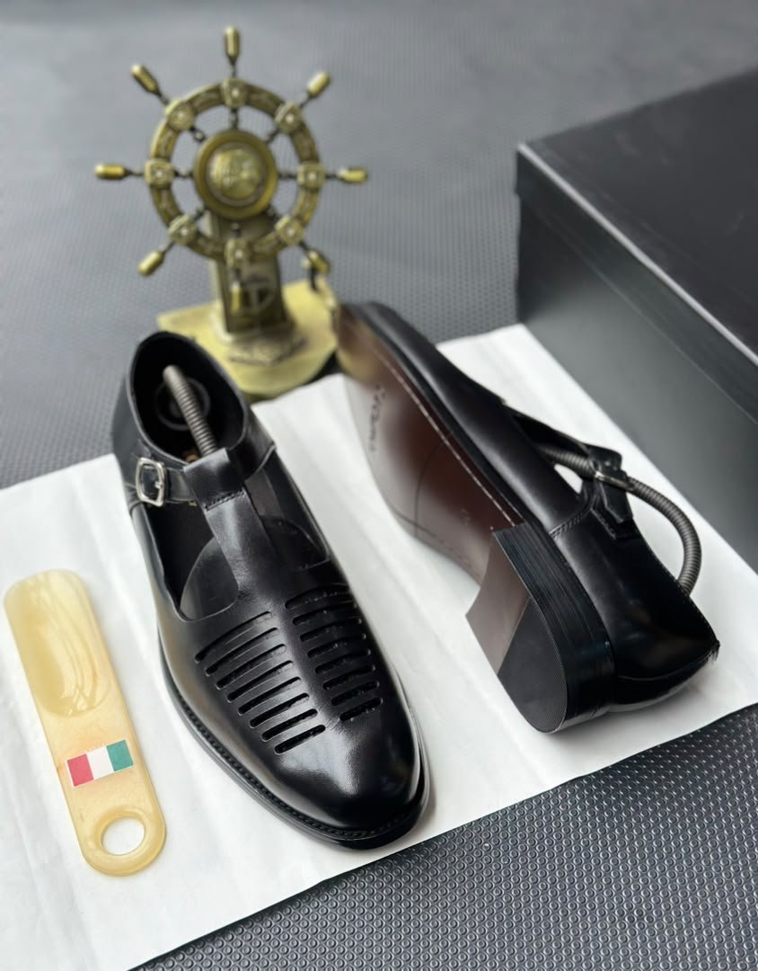root A pair of luxurious black leather shoes with a silver buckle and ventilation details, presented on a white cloth with a shoe tree and a decorative gold ship's wheel in the background.