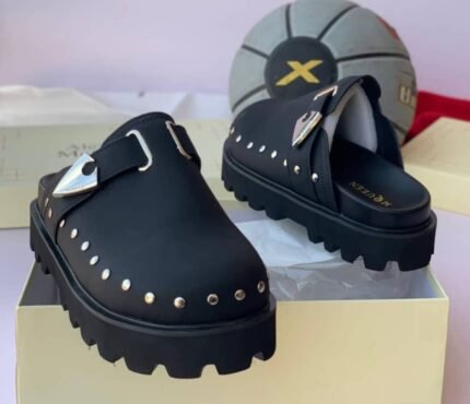 A pair of luxurious green half shoes with metallic studs and a sophisticated buckle, showcased on a cream-colored shoe box.