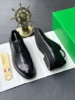 A pair of luxurious black leather oxfords showcased on a crisp white surface, accompanied by a shoe tree and a green box, exuding an air of sophistication.