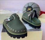 A pair of luxurious green half shoes with metallic studs and a sophisticated buckle, showcased on a cream-colored shoe box.