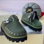A pair of luxurious green half shoes with metallic studs and a sophisticated buckle, showcased on a cream-colored shoe box.