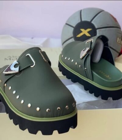 A pair of luxurious green half shoes with metallic studs and a sophisticated buckle, showcased on a cream-colored shoe box.