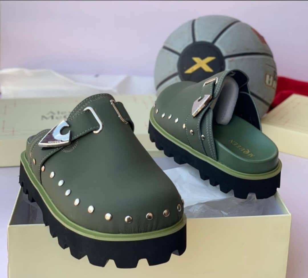 root A pair of luxurious green half shoes with metallic studs and a sophisticated buckle, showcased on a cream-colored shoe box.