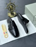 A pair of luxurious black leather half shoes displayed on a white surface, accompanied by a small Italian flag and a decorative gold ship's wheel, exuding elegance and refinement.