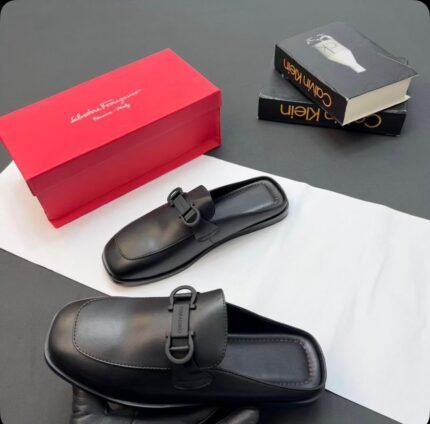 A pair of luxurious black leather half shoes with a sleek buckle, accompanied by a red shoe box and two fashion books, set against a sophisticated gray and white background.