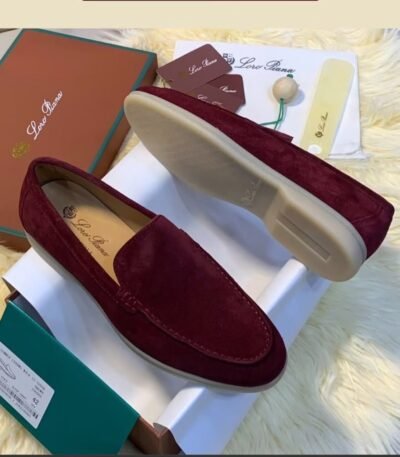 A pair of luxurious maroon loafers with velvety texture, displayed on a white shoe box with accessories and packaging in the background.