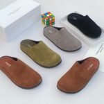 A stylish arrangement of four suede half shoes in various earthy tones, accompanied by their elegant white boxes and a vibrant Rubik's Cube, set against a pristine white backdrop.