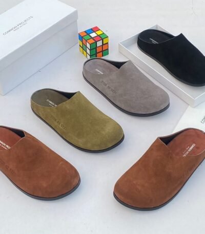 A stylish arrangement of four suede half shoes in various earthy tones, accompanied by their elegant white boxes and a vibrant Rubik's Cube, set against a pristine white backdrop.