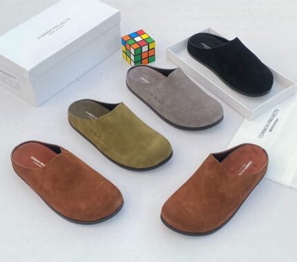 A stylish arrangement of four suede half shoes in various earthy tones, accompanied by their elegant white boxes and a vibrant Rubik's Cube, set against a pristine white backdrop.